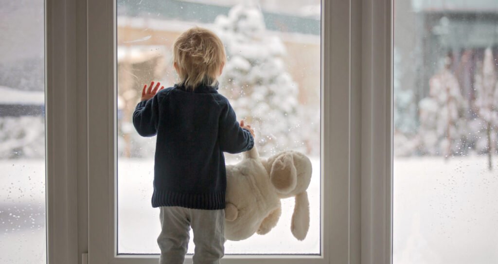 Toddler child standing in front of a big french doors, leaning against it looking outside at a snowy nature, holding teddy toy
