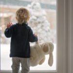 Toddler child standing in front of a big french doors, leaning against it looking outside at a snowy nature, holding teddy toy